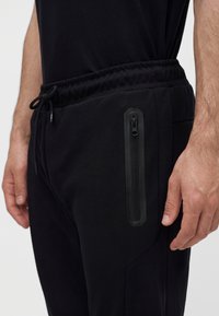 Gros plan d'une personne portant un jogger noir avec une taille élastique et une poche verticale zippée imperméable sur la cuisse droite.