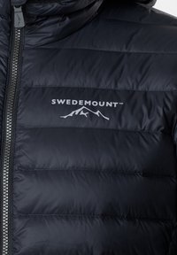 Giacca imbottita blu navy con cuciture orizzontali, zip frontale e logo Swedemount bianco. Materiale liscio e lucido con dettaglio del cappuccio.