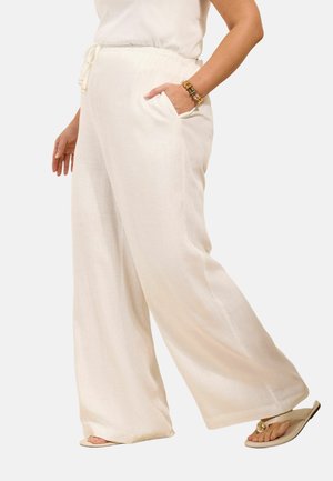 Kobieta w luźnych białych spodniach z wysokim stanem i kieszeniami, nosząca biały top, beżowe sandały i koralikową bransoletkę na nadgarstku.