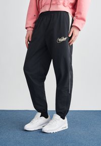Μαύροι παντελόνια Nike joggers με ελαστική μέση, λευκές πλαϊνές ρίγες και χρυσό λογότυπο. Συνδυασμένα με λευκά αθλητικά παπούτσια Air Max σε μπλε χαλί.