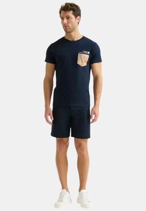 T-shirt blu navy con tasca marrone chiaro decorata con una mappa. Abbinato a pantaloni corti blu navy e sneaker bianche. Maniche corte, scollatura tonda.