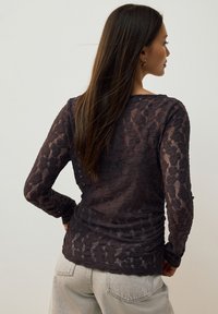 Dlouhý rukáv topu v tmavě hnědé barvě s texturovaným krajkovým vzorem. Látka působí lehce a má přiléhavý střih.