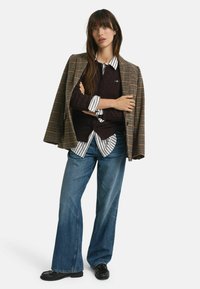 Braun karierter Blazer über einem dunklen Strickpullover, gestreiftem Hemd in Weiß und Schwarz, ausgestellten blauen Jeans und schwarzen Schuhen. Lange Haare, neutraler Hintergrund.