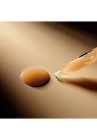 Bouteille compte-gouttes en verre à côté d'une goutte de liquide beige clair sur un fond neutre, mettant en avant la texture lisse et le subtil éclat.