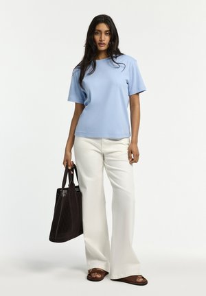 Frau mit langen dunklen Haaren, die ein hellblaues kurzärmeliges Oberteil, weiße Hosen mit weitem Bein, braune Sandalen trägt und eine große dunkle Tragetasche hält.