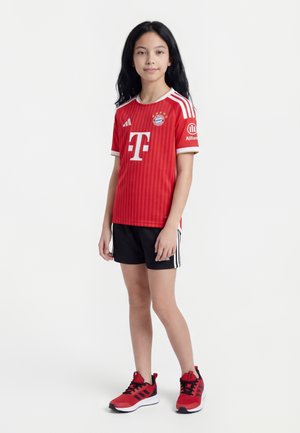 Dziewczyna stoi, ubrana w czerwoną koszulkę piłkarską Bayernu Monachium, czarne spodenki Adidas i czerwone buty sportowe Adidas na białym tle.