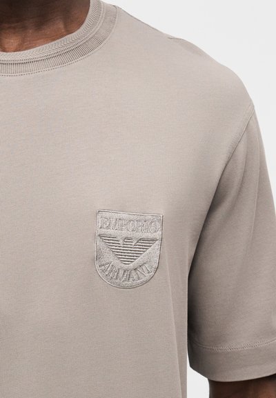 Light gray short-sleeve t-shirt made of soft fabric, featuring a textured embroidered logo on the left chest with a shield design.