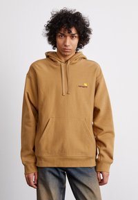 Brown hooded sweatshirt with a front pocket, ribbed cuffs, and a small logo on the left chest. Material appears soft and fleece-like.