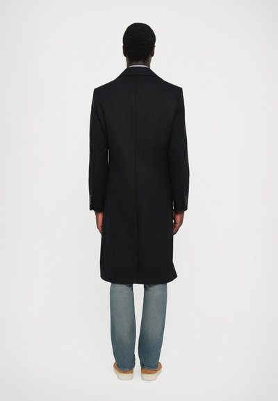 Palton negru cu un design pe măsură, siluetă lungă și textură netedă. Purtați peste blugi de culoare albastru deschis și pantofi bej, văzuți din spate.