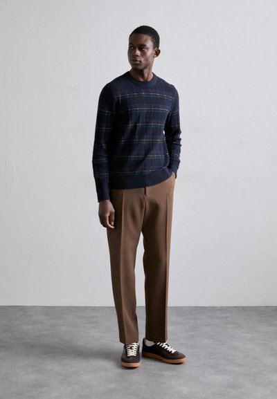 Pullover azul-marinho com padrões xadrez verde e amarelo, combinado com calças castanhas ajustadas e sapatilhas escuras com atacadores brancos. Texturas de tecido suaves visíveis.