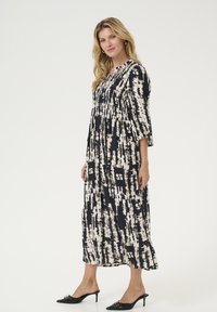 Robe fluide noire et crème avec un motif abstrait, corsage smocké, manches trois-quarts, et ourlet longueur cheville. Associée à des mules à talons noirs.