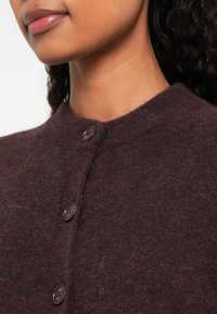 Mulher vestindo um cardigan felpudo botão escuro roxo, mostrando o pescoço e a parte inferior do rosto com os lábios fechados.