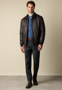 Muž oblečený v tmavě hnědé kožené bundě, modré denimové košili, béžovém roláku, černých kalhotách, hnědém pásku a tmavě hnědých botách stojící na šedé podlaze.