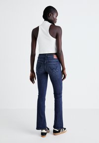 Débardeur blanc court avec col rond, associé à un jean évasé bleu foncé avec poches arrière et un patch en cuir marron.