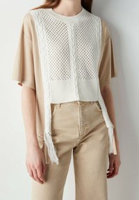 Donna che indossa pantaloni beige e una maglietta beige a maniche corte larga, con un gilet senza maniche bianco lavorato a maglia, decorato con frange.