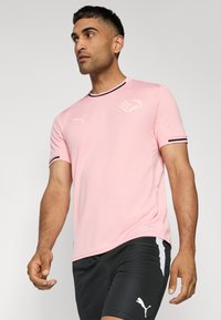 T-shirt de sport rose clair à manches courtes, avec des accents noirs sur le col et les manches, présentant un logo blanc sur la poitrine.