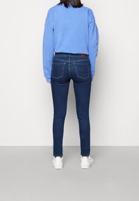 Sudadera azul, jeans ajustados de mezclilla oscura, vista trasera, con parche de cuero de la marca en la cintura y zapatillas blancas. Textura de tela suave.