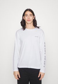 T-shirt de manga longa branca feita de algodão, com um pequeno logótipo no peito e números de coordenadas impressos ao longo da manga esquerda.