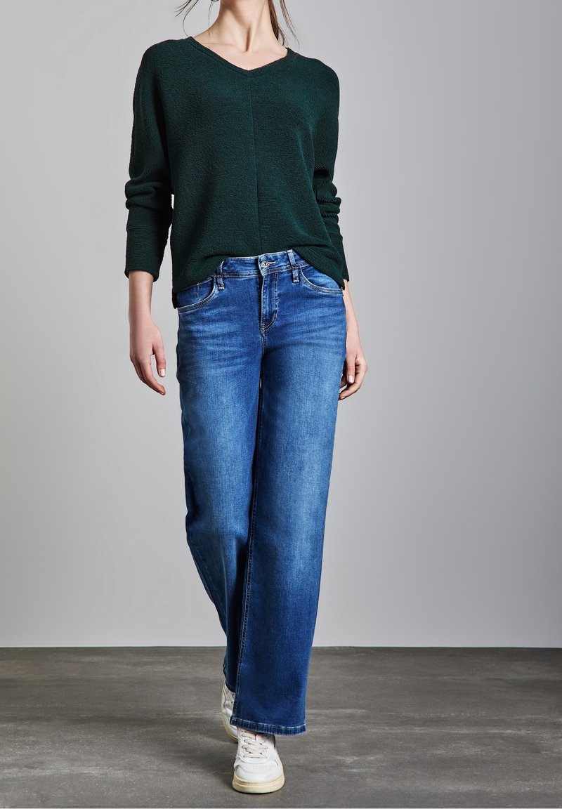 Maglione verde scuro con tessitura e scollo a V, abbinato a jeans a gamba larga di colore azzurro chiaro. Il modello indossa sneakers bianche. Design semplice e moderno.