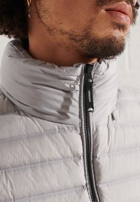 Veste en duvet gris clair avec col haut, texture matelassée horizontale, fermeture éclair à l'avant et détail de tirette de fermeture à glissière avec logo.