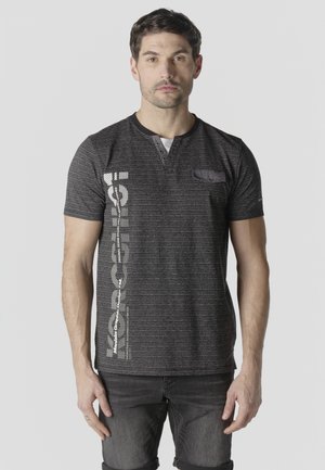 Muž stojaci čelom vpredu, oblečený v čierno-šedej pruhovanej tričku s krátkym rukávom a bielym vertikálnym textom na ľavej strane.