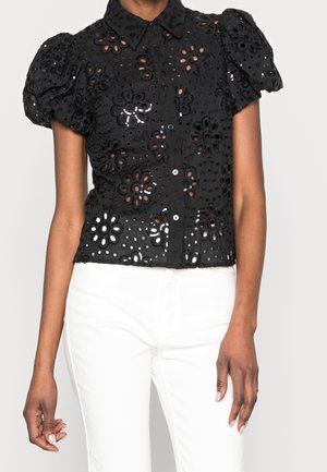 Vrouw draagt een zwarte blouse met korte mouwen, bloemenopeningen en pofmouwen, gecombineerd met een witte broek met hoge taille.
