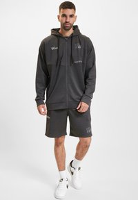 Felpa grigia con zip, cappuccio regolabile e dettagli con logo, abbinata a pantaloni corti coordinati, presenta una vestibilità comoda e una texture morbida, indossata con sneaker bianche.