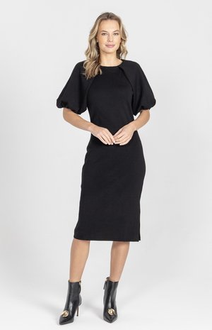 Kobieta stojąca z rękami splecionymi, ubrana w czarną sukienkę midi z bufkami i czarne botki na obcasie ze złotymi akcentami.