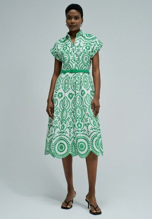 Mujer con vestido midi blanco con patrones circulares verdes, mangas cortas, cinturón verde y sandalias negras de tacón, de pie frente a un fondo liso.