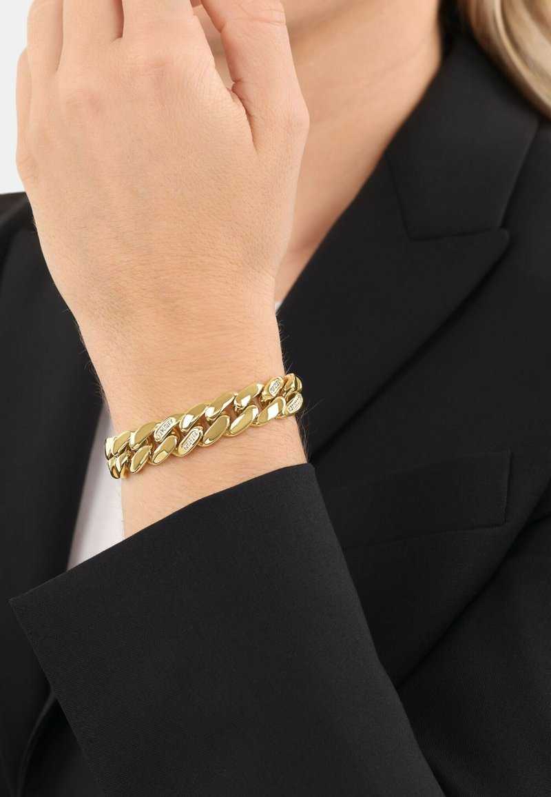 Muñeca con una pulsera gruesa de cadena dorada brillante que presenta pequeños diamantes incrustados, llevada sobre la manga de una chaqueta negra.