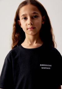 Jeune fille aux longs cheveux bruns portant un t-shirt noir "American Vintage", tournée vers l'avant avec une expression neutre sur un fond uni.