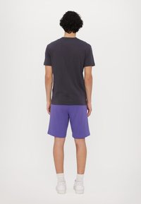 Short-sleeved dark gray t-shirt with a crew neck, paired with purple shorts. The fabric appears soft with a loose fit. White sneakers are worn.