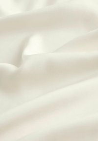 Bílá tkanina s hladkým, mírně texturovaným povrchem, zavěšená v jemných záhybech, ukazující subtilní odrazy světla a lehký lesk.