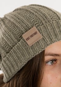 Gebreide muts in gedempt groen, met een gestructureerd ribbelontwerp en een bruine leren label met de tekst "Knit Factory" aan de zijkant.
