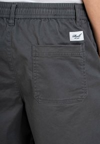 Graue Shorts mit elastischem Bund, die eine große Gesäßtasche mit einem Markenlabel aufweisen. Der Stoff hat eine glatte, matte Textur und aufgenähte Details.