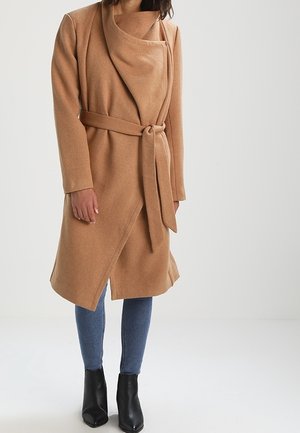 Vrouw draagt een beige wikkeljas tot op de knie, gestrikt in de taille, blauwe skinny jeans en zwarte enkellaarsjes, staand tegen een effen achtergrond.