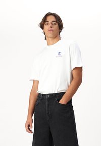 Hvit bomulls-T-skjorte med korte ermer, med en blå trykt logo på brystet. Kombinert med svarte denim-bukser, teksturert med et slitt utseende.