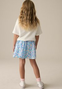 Chemise blanche à manches courtes associée à une jupe fleurie colorée et scintillante bleu et rose. Baskets montantes blanches avec des chaussettes roses. Cheveux texturés.