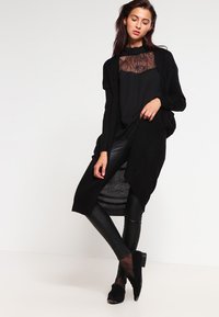 Zwarte gebreide cardigan met een open voorkant, gelaagd over een doorschijnende zwarte kanten top en slim-fit zwarte leggings, gecombineerd met zwarte instappers.