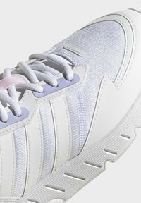 Zapatilla blanca con parte superior texturizada, que presenta acentos en púrpura claro, cordones planos y un patrón de suela ondulada para mayor tracción.