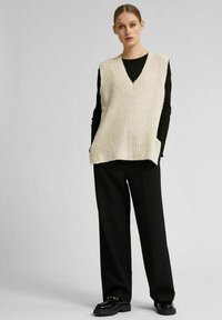 Gilet en tricot beige avec un décolleté en V profond, porté sur un haut à manches longues noir. Présente une coupe ample et des poches latérales, associé à un pantalon noir large.