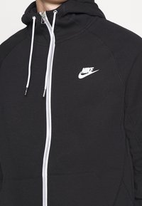 Černá mikina na zip vyrobená z měkké látky, s bílým logem Nike na levé straně hrudi, kapucí se stahovací šňůrkou a předními kapsami.