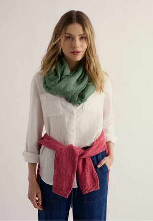 Vrouw met los golvend haar, die een wit overhemd met knopen draagt, een groene sjaal, blauwe spijkerbroek en een roze trui om haar middel geknoopt.