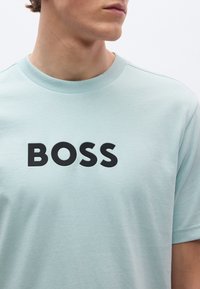 Hele sinine puuvillane t-särk ümara kaelusega, millel on rinnal julge must "BOSS" logo. Sile tekstuur, lühikesed varrukad.