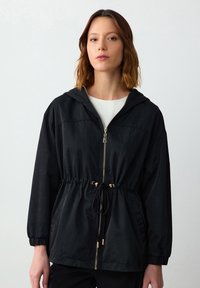 Zwarte lichtgewicht jas met een capuchon, aangehaalde taille, gouden hardware, elastische manchetten en een gladde textuur. Voorzien van een ritssluiting aan de voorkant.