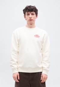 Νεαρός άνδρας με σκούρα σγουρά μαλλιά, που φοράει μπεζ φούτερ Levi Strauss και καφέ παντελόνι, στέκεται μπροστά σε απλό φωτεινό φόντο.