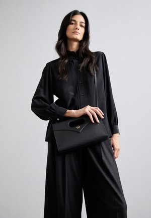 Schwarze strukturierte Handtasche mit einem oberen Griff, strukturiertem Leder und goldenen Hardware-Akzenten. Modell trägt eine schwarze, plissierte Bluse und Hosen.