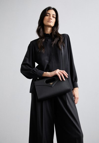Sac à main noir structuré avec une poignée supérieure, en cuir texturé et accents en métal doré. Modèle portant une blouse plissée noire et un pantalon.