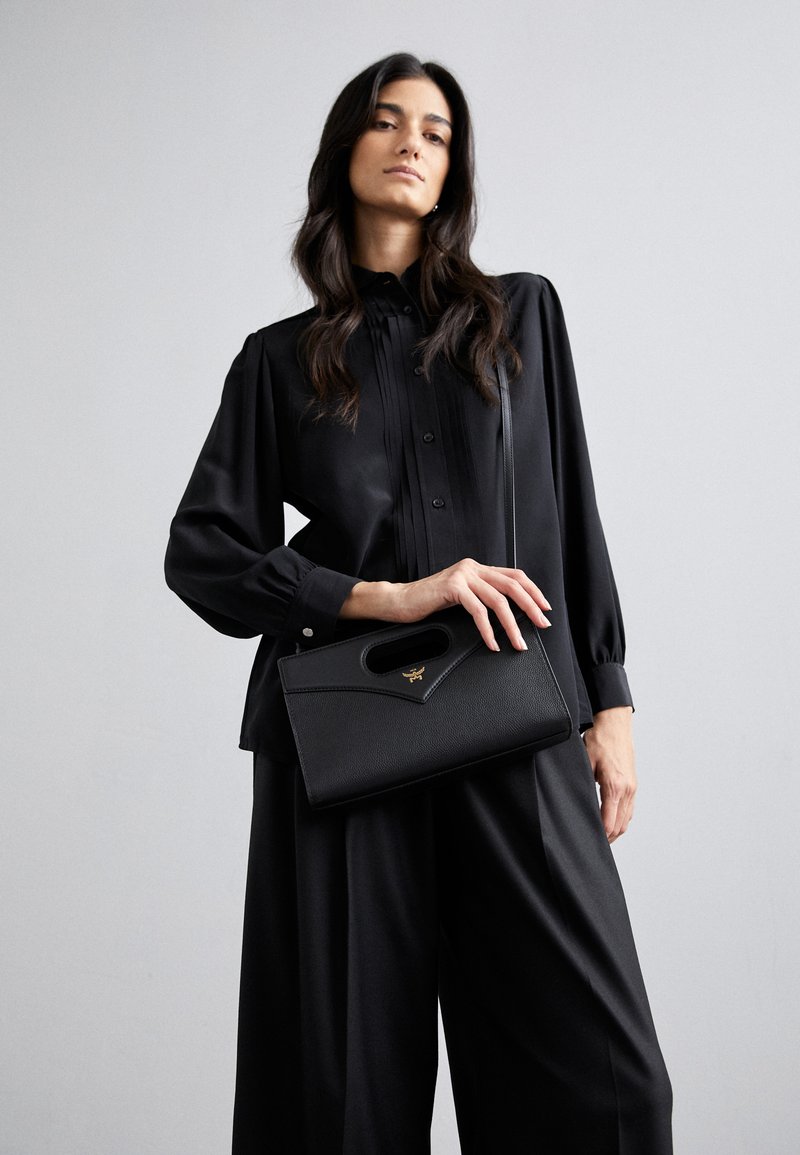 Sorte struktureret håndtaske med et top håndtag, tekstureret læder og guld hardware accents. Model iført en sort, plisseret bluse og bukser.