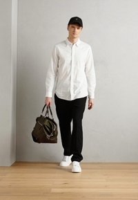 White button-up shirt, black pants, and white sneakers. Carrying a green bag with gray accents and hardware. Background is light gray.